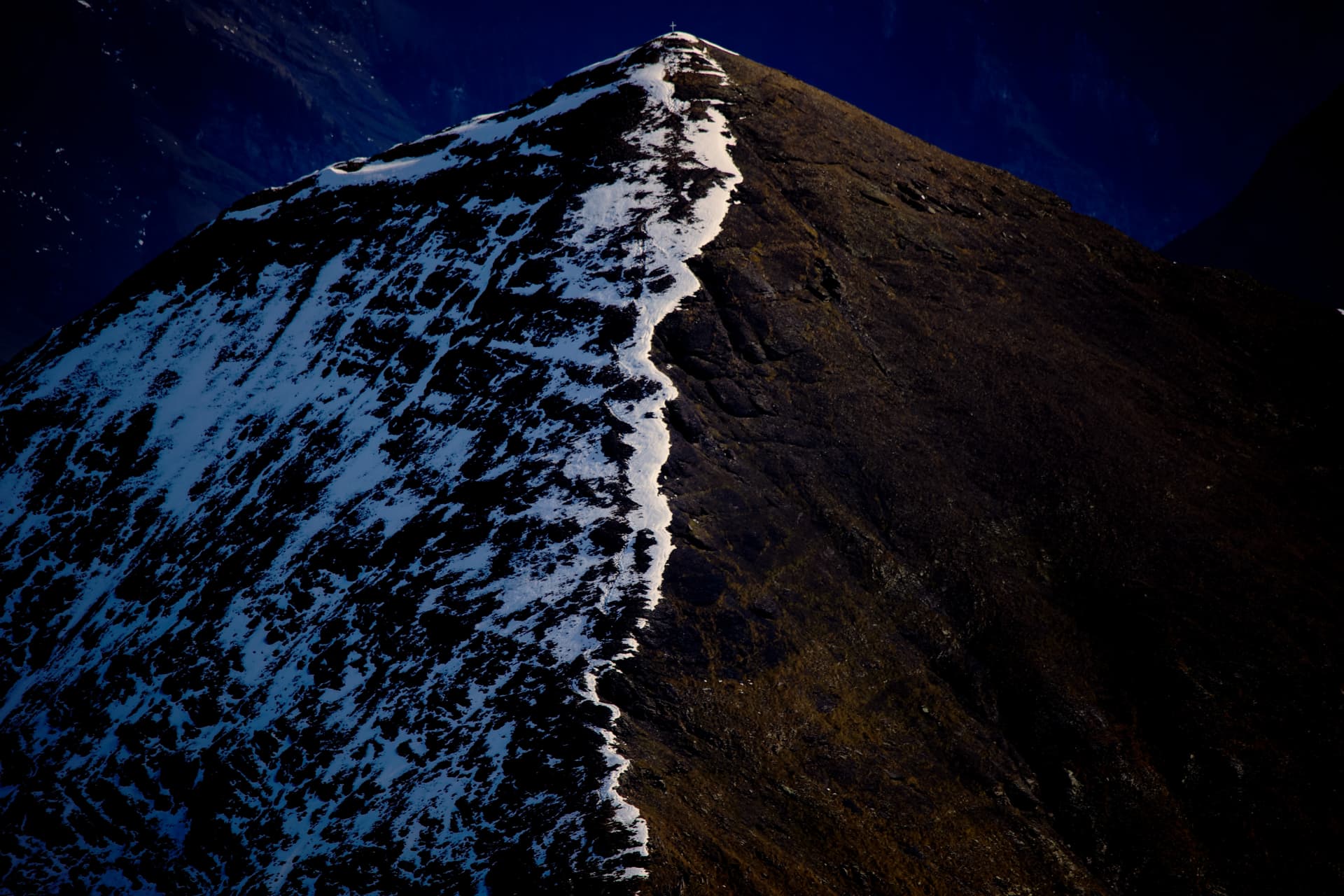 Scharfer Grat mit Gipfelkreuz und Schneeband im blauen Schatten