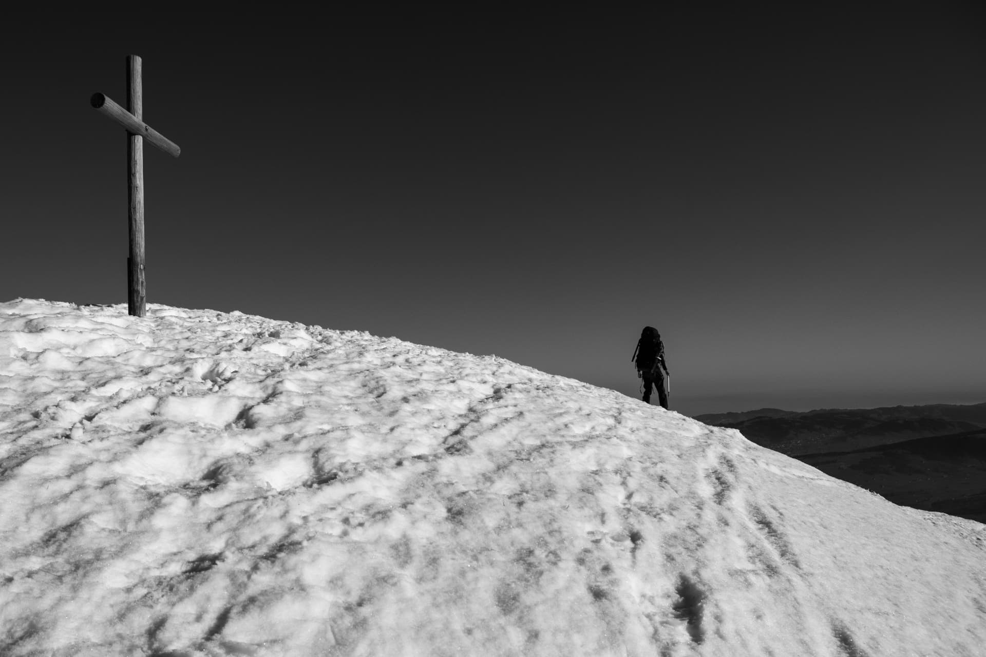 Schwarz-Weiß-Foto eines Holzkreuzes auf einem verschneiten Gipfel mit der Silhouette eines Wanderers