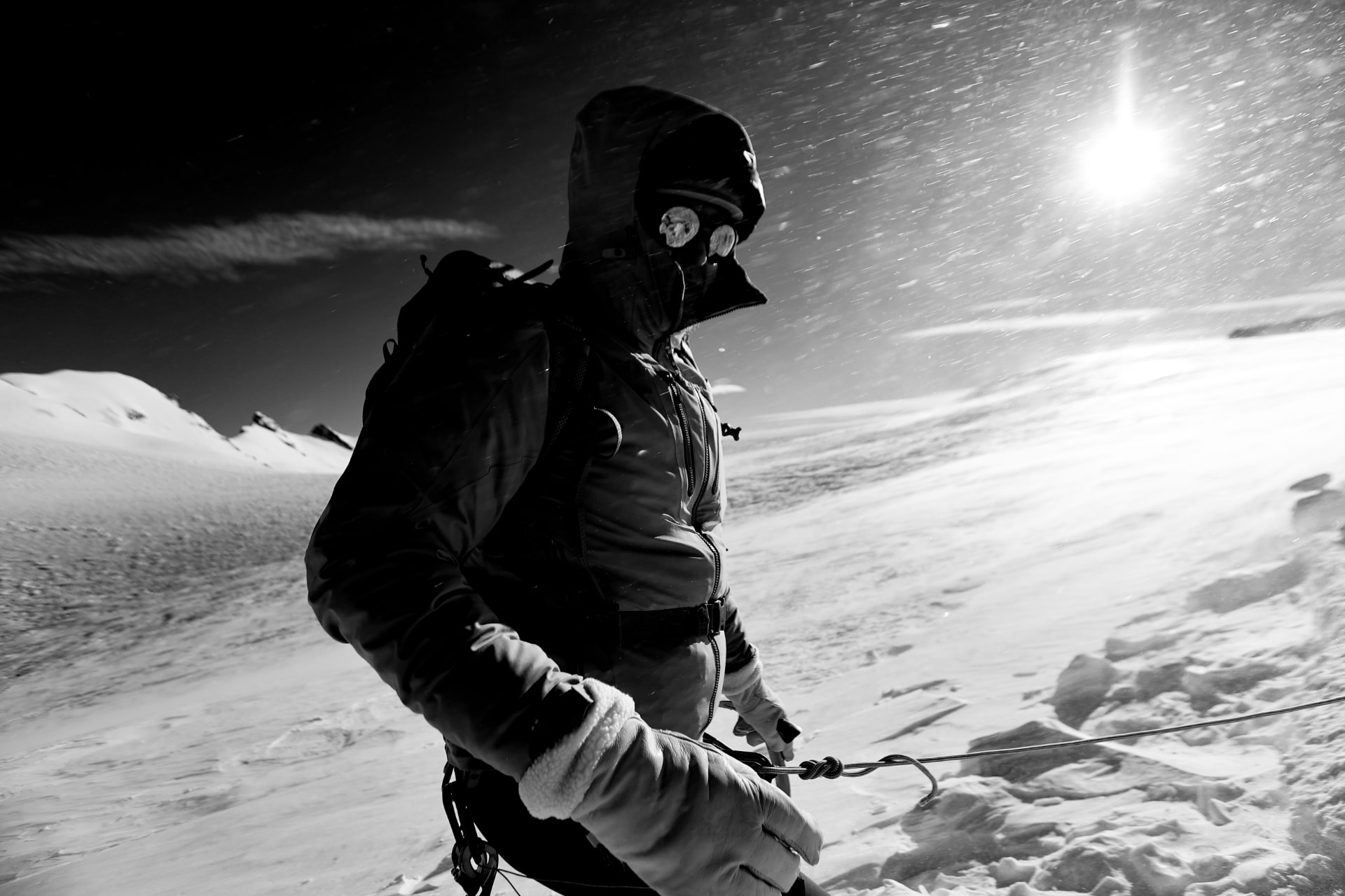 Bergsteiger mit Kapuze und Brille im Schneetreiben, tiefe Sonne über dem Gletscher, schwarzweiss