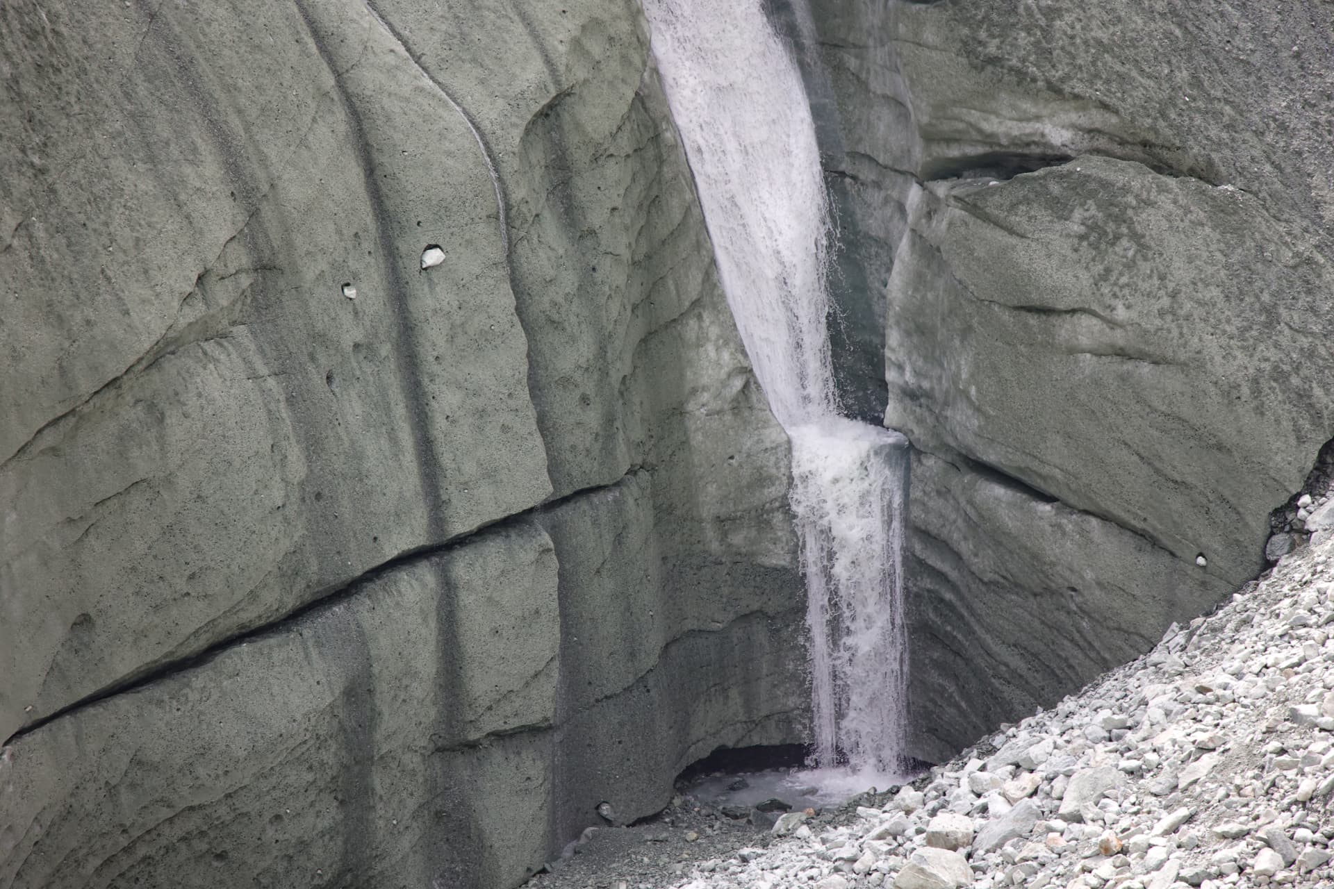 Schmelzwasserfall über eine Gletscherwand in grauem Schutt
