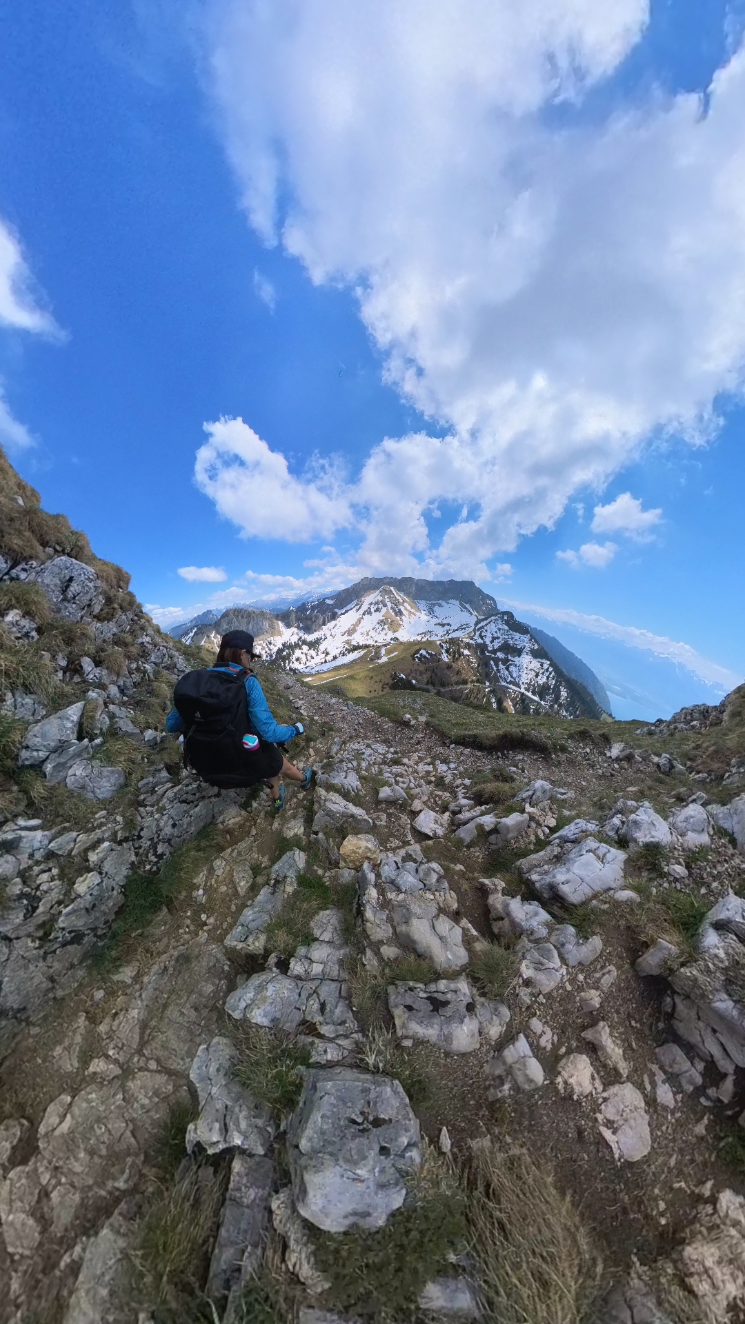 Wanderin in blauer Jacke ruht auf einem steinigen Weg unter einem schneebedeckten Grat